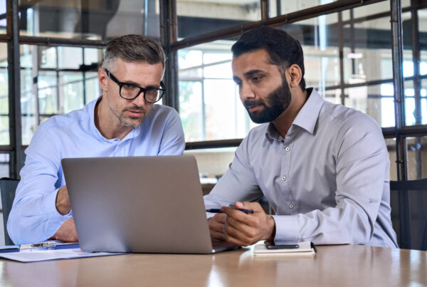 2 hommes discutent, assis à un bureau, derrière un ordinateur portable