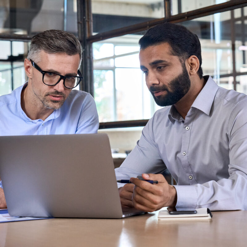 2 hommes discutent, assis à un bureau, derrière un ordinateur portable